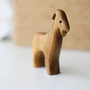 May include: A handcrafted wooden goat figurine. The goat is carved from light brown wood, with a simple, stylized design. It has a small, rounded body, short legs, and a slightly curved horn. The figurine is standing on a white surface.