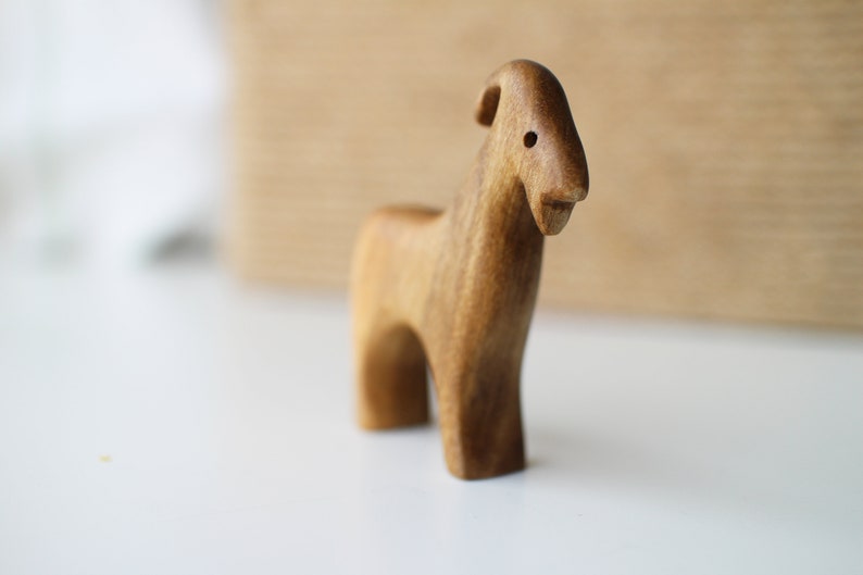 May include: A handcrafted wooden goat figurine. The goat is carved from light brown wood, with a simple, stylized design. It has a small, rounded body, short legs, and a slightly curved horn. The figurine is standing on a white surface.