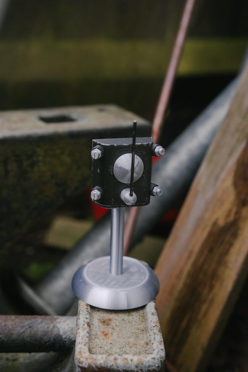 May include: A small, black, rectangular device with a silver circular element and a thin, vertical rod. The device is mounted on a silver stand with a circular base. The object is secured with silver bolts.