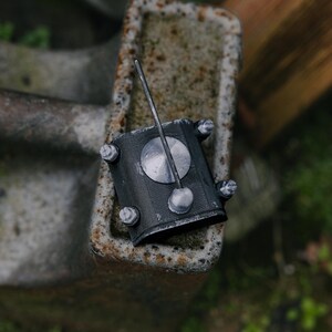 May include: A dark grey metal object with a circular top and a long, thin rod extending upwards. The object is attached to a rectangular base with bolts. The background is blurred, with hints of green foliage and weathered surfaces.