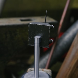 May include: A small, handmade metal sculpture. It has a black, cube-shaped top with a thin antenna and two small, cylindrical attachments. It is mounted on a silver, cylindrical post and a circular base. The background is blurred.