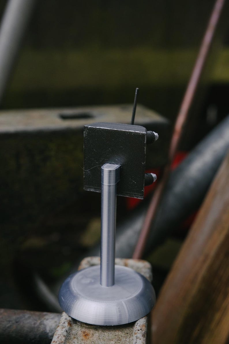 May include: A small, handmade metal sculpture. It has a black, cube-shaped top with a thin antenna and two small, cylindrical attachments. It is mounted on a silver, cylindrical post and a circular base. The background is blurred.