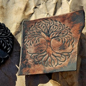 Puede incluir: Dos objetos decorativos con un diseño de árbol de la vida. Uno es una pieza negra y circular con un detallado tallado de árbol. El otro es una pieza rectangular marrón con un diseño de árbol similar, que muestra una estética rústica.