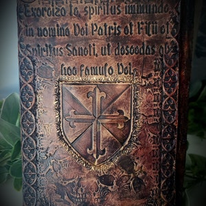May include: A leather-bound book with a gold embossed design. The cover features a shield with a cross and the text "Exorcizo to, spiritus immundo, in nomino Dei Patris of Filii of Spiritus Sanoti, ut desoodas obs froo famulo Dei."