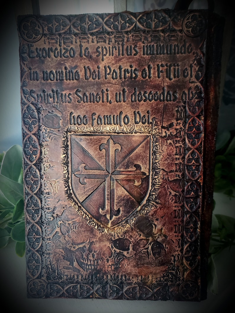 May include: A leather-bound book with a gold embossed design. The cover features a shield with a cross and the text "Exorcizo to, spiritus immundo, in nomino Dei Patris of Filii of Spiritus Sanoti, ut desoodas obs froo famulo Dei."