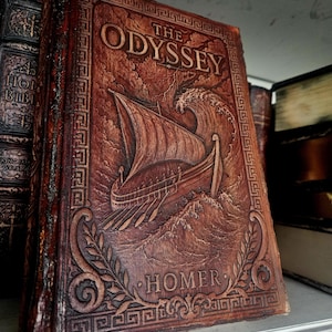 Puede incluir: Un libro encuadernado en cuero marrón titulado The Odyssey de Homer. La portada presenta una ilustración en relieve de un barco antiguo navegando entre olas turbulentas bajo un cielo tormentoso, enmarcado por un borde con patrón griego.