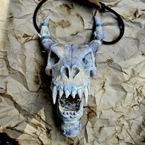 Può includere: Ciondolo a forma di teschio di drago dipinto a mano con cordino nero. Il teschio presenta sfumature blu, viola e gialle, con denti e corna affilate. Adatto per appassionati di cosplay o fantasy.