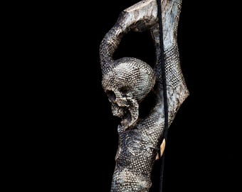 Może przedstawiać: Laska z wyrzeźbioną czaszką zintegrowaną z teksturowaną, przypominającą gałąź strukturą o metalicznym, postarzanym wykończeniu. Powierzchnia wykazuje skomplikowane, łuskopodobne wzory, a z górnej części zwisa cienki czarny sznurek.
