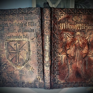 May include: A leather-bound book with a worn, aged appearance. The cover features a gold-colored cross and the text "Exorcizo to spiritus immundo. In nomine Doi Patris of Filii of Snielius Sanoti, ut descodas ob fioo famulo Deis". The other side of the book has the text "The Mallus Maleficarum" and a detailed illustration of a figure in a robe.