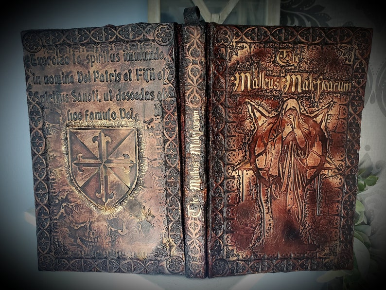 May include: A leather-bound book with a worn, aged appearance. The cover features a gold-colored cross and the text "Exorcizo to spiritus immundo. In nomine Doi Patris of Filii of Snielius Sanoti, ut descodas ob fioo famulo Deis". The other side of the book has the text "The Mallus Maleficarum" and a detailed illustration of a figure in a robe.
