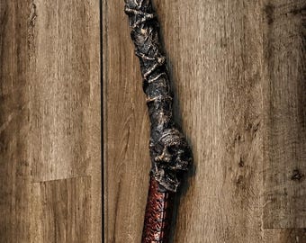 May include: A long, curved bow featuring a dark, textured upper limb and a reddish-brown, stitched leather-like grip. The bow is adorned with two sculpted skull motifs and intricate, gnarled branch-like detailing along the frame.