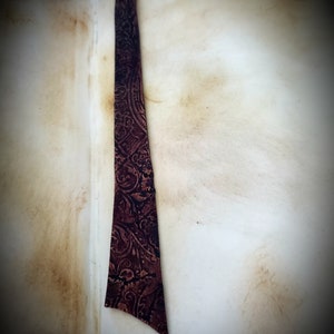 May include: A close-up of a brown leather bookmark with a paisley pattern. The bookmark is attached to a book with a worn, cream-colored cover.