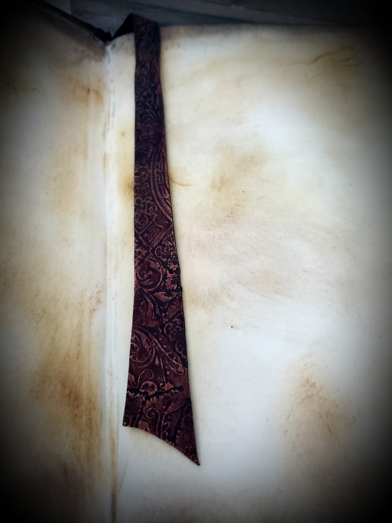 May include: A close-up of a brown leather bookmark with a paisley pattern. The bookmark is attached to a book with a worn, cream-colored cover.