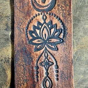 May include: A brown leather item with a detailed black embossed design. The design includes a crescent moon, a lotus flower, and other geometric shapes. The leather has a textured, aged appearance.