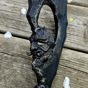 Puede incluir: Una escultura negra y ornamentada de una máscara de demonio japonés con un largo bigote curvado. La máscara está detallada con rasgos texturizados y se encuentra sobre un fondo de madera con pétalos blancos dispersos.