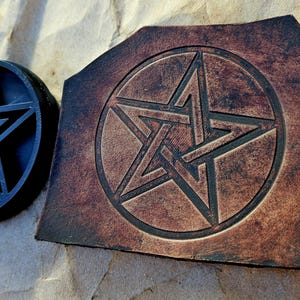 May include: A black, circular stamp and a brown leather piece, both featuring a pentagram design. The stamp is raised, while the leather piece has an etched design. The items are displayed on a textured, brown surface.
