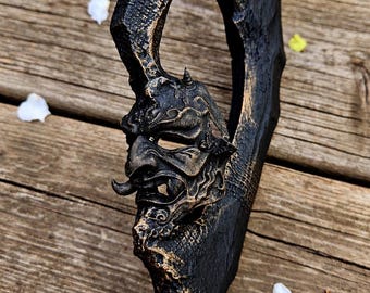 May include: A black staff featuring a sculpted Hannya mask at the top, finished with gold-toned highlights. The handle has a textured, rugged appearance with a curved, open-work design above the mask, showcasing intricate detailing.