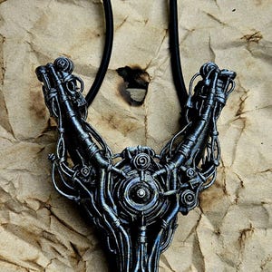 Può includere: Una fionda in stile futuristico con un design metallico dettagliato. La fionda presenta una complessa rete di fili ed elementi meccanici, con un sacchetto di pelle marrone e fasce nere. Lo sfondo è una carta marrone strutturata.