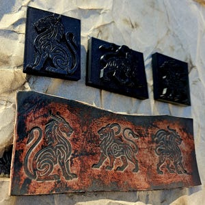 Può includere: Una collezione di sculture a rilievo nere di creature mitiche, tra cui leoni e un drago, montate su un muro di pietra strutturato. Le sculture sono rettangolari e quadrate, con dettagli intricati. Un pezzo più grande, marrone, simile alla pelle, presenta ulteriori sculture.