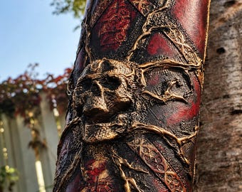 Könnte beinhalten: Ein zylindrisches Objekt aus Leder mit tiefroter Oberfläche, komplexen geprägten geometrischen Mustern und einer markanten dreidimensionalen Totenkopf-Skulptur in der Mitte, umgeben von erhabenen, astartigen goldenen Details.