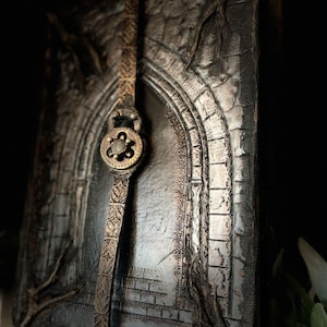 May include: A black leather journal with a silver and gold embossed design of an archway. The journal has a leather strap closure with a decorative metal clasp.