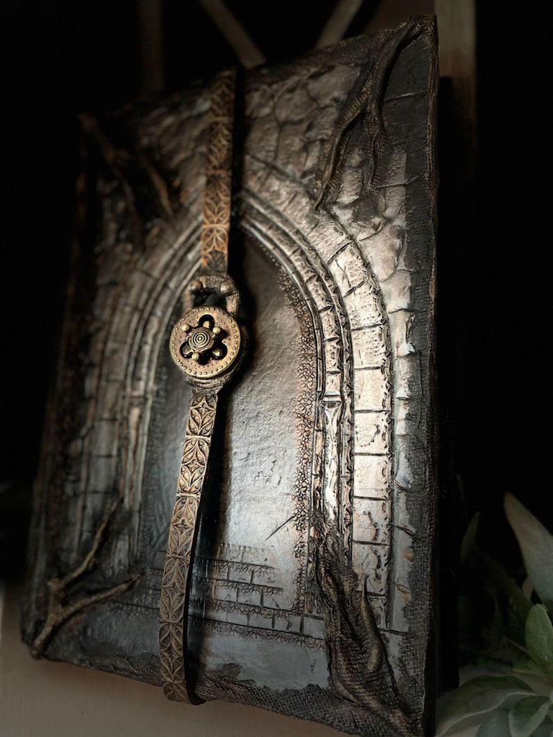 May include: A black leather journal with a silver and gold embossed design of an archway. The journal has a leather strap closure with a decorative metal clasp.