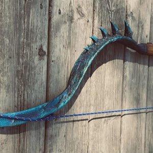 Könnte beinhalten: Handgefertigter Bogen mit einzigartigem, geschwungenem Design. Der Bogen hat eine strukturierte, türkisfarbene Oberfläche mit dunklen Akzenten und einem Holzgriff. Eine blaue Schnur ist über den Bogen gespannt. Der Bogen ist auf einer verwitterten Holzoberfläche montiert.