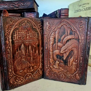 May include: Two brown leather-bound books with intricate embossed covers. The left book features the title Harry Potter and the Philosopher's Stone with a castle illustration. The right book depicts a figure facing a large serpent in a stone chamber.