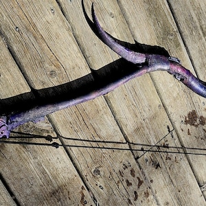Può includere: Un arco curvo di colore viola con intricati motivi intagliati a forma di foglia sul flettente inferiore e protuberanze appuntite simili a corna sulla sezione superiore. L'arco è teso con un sottile cordino nero tra le due estremità.