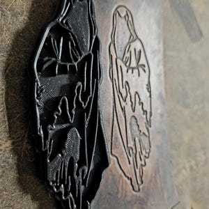 May include: A black 3D-printed stamp of a hooded figure, next to an embossed image of the same figure on a piece of leather. The stamp is a solid, raised relief, while the leather image is a recessed impression. The leather has a distressed, aged appearance.
