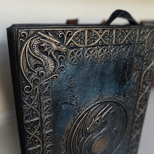 May include: A dark blue, embossed book with a dragon design. The cover features a detailed dragon head in the upper left corner and a circular dragon emblem in the center. The edges and details are highlighted in gold.