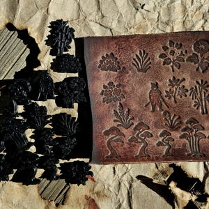 Puede incluir: Un cuadrado de cuero marrón con diseños botánicos y figuras japonesas en relieve. Junto a él, varias formas negras recortadas de árboles y follaje, todo sobre un fondo de papel beige texturizado.