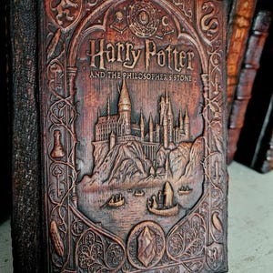 May include: A brown, carved wooden book cover titled Harry Potter and the Philosopher's Stone by J.K. Rowling. The cover features a detailed relief of a castle on a cliff, small boats on water, and intricate border designs including a lion and snake.