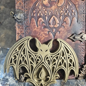 May include: A gold-coloured bat-shaped embellishment sits atop a brown, textured surface, possibly part of a book. The surface has a matching embossed bat design. The overall style is gothic, with intricate detailing on the bat and the surrounding elements.
