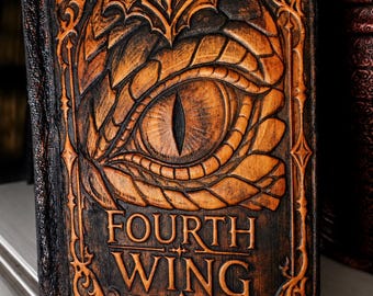 May include: A rectangular book with a dark brown, textured cover featuring a carved dragon eye at the top and the text "FOURTH WING" embossed in the center. The cover is decorated with intricate, raised scrollwork and border patterns.