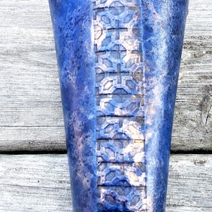 Puede incluir: Un jarrón de cerámica azul con un patrón geométrico y un borde decorativo. El jarrón tiene un acabado desgastado y un brillo metálico.