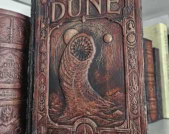 以下が含まれることがあります： DUNEというタイトルが浮き彫りになった、ダークブラウンの質感のある表紙を持つ長方形の本。表紙には、巨大なサンドワーム、天体、そしてナイフのアイコンを含む複雑な縁取りの彫刻が中央のレリーフとして施されています。