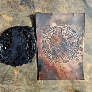 May include: A black, circular emblem and a rectangular leather piece with a Grim Reaper design. The emblem is 3D printed. The leather piece features a Grim Reaper with a scythe in front of a clock face.