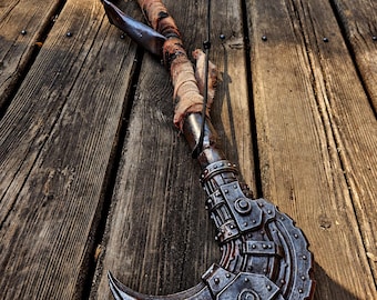 May include: A long, curved prop weapon featuring a metallic, dark grey axe-like head with visible rivets and mechanical detailing. The handle is wrapped in weathered, frayed tan and brown fabric strips, leading to a dark, textured grip at the end.
