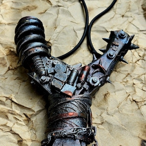 Può includere: Una fionda artigianale con un'estetica steampunk. La fionda presenta un telaio in metallo con dettagli intricati, tra cui un'impugnatura a spirale, giunti rivettati e una testa a punte. La sacca in pelle è attaccata con un tubo di gomma nero.
