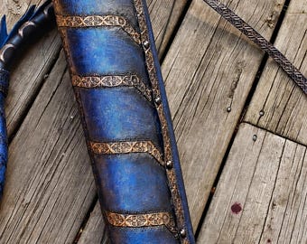 Könnte beinhalten: Ein zylindrischer blauer Köcher mit strukturierter Oberfläche, verziert mit bronzefarbenen Bändern und komplexen geometrischen Mustern. Der Köcher ist seitlich mit Metallnieten befestigt und verfügt über einen passenden gemusterten Riemen.