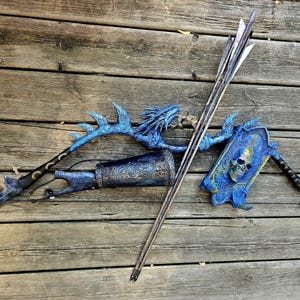 Könnte beinhalten: Ein Fantasy-Bogen- und Pfeil-Set mit blauem Drachendesign. Der Bogen weist aufwendige Details auf und ist mit einem Köcher mit Pfeilen kombiniert. Das Set befindet sich auf einer Holzoberfläche.