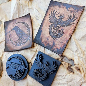 May include: Three leather stamps with different designs. One stamp features a raven perched on a crescent moon, another features a phoenix with its wings spread, and the third features a phoenix with its wings spread in a more stylized design.