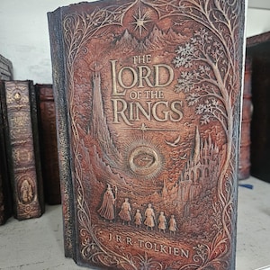Op de afbeelding: Een gebonden boek getiteld The Lord of the Rings door J.R.R. Tolkien met een bruine, in reliëf gemaakte kaft. De voorzijde toont een gedetailleerde reliëfafbeelding van een toren, een ring en een groep figuren, omlijst door boom- en rankmotieven.