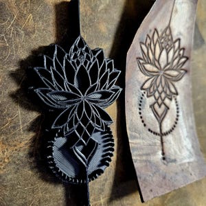 May include: A black lotus flower stamp and its impression on leather. The 3D-printed stamp displays detailed petals and a decorative base. The leather has a brown, textured appearance. The impression is a mirror image of the stamp.