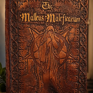 Puede incluir: Un libro rectangular con una cubierta de color marrón oscuro texturizada que presenta un pentagrama en relieve y una figura central. El título The Malleus Maleficarum está impreso en letras realzadas en la parte superior, rodeado por un borde complejo.