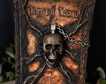 May include: A rectangular book with a textured, dark brown and copper-colored cover featuring the embossed text Tome of Tears. A three-dimensional skull is centered on the front, connected to a silver-toned metal chain link.