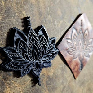 May include: A black lotus flower-shaped stamp and a leather piece with an embossed lotus design. The stamp is 3D-printed with intricate details. The leather piece is diamond-shaped with a brown hue, showcasing the embossed floral pattern.