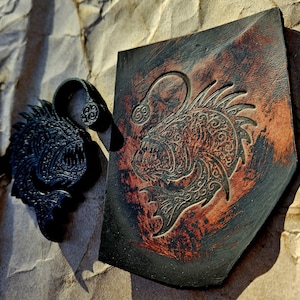 May include: A black, textured, fish-shaped pendant and a leather shield with a fish design. The shield is brown and black with a carved fish and the number 69. The pendant has a matching fish design.
