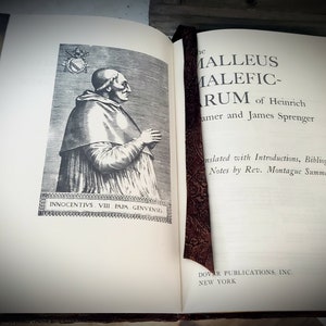 May include: An open book with a brown leather cover and a bookmark. The book is titled "The Malleus Maleficarum" by Heinrich Kramer and James Sprenger. The book is translated with introductions, bibliography, and notes by Rev. Montague Summers. Dover Publications, Inc. New York.